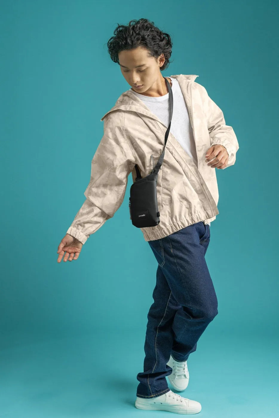 Pacsafe Go Anti-Theft Tech Crossbody Bag in Black, worn crossbody over a light jacket against a teal backdrop