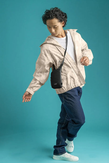Pacsafe Go Anti-Theft Tech Crossbody Bag in Black, worn crossbody over a light jacket against a teal backdrop