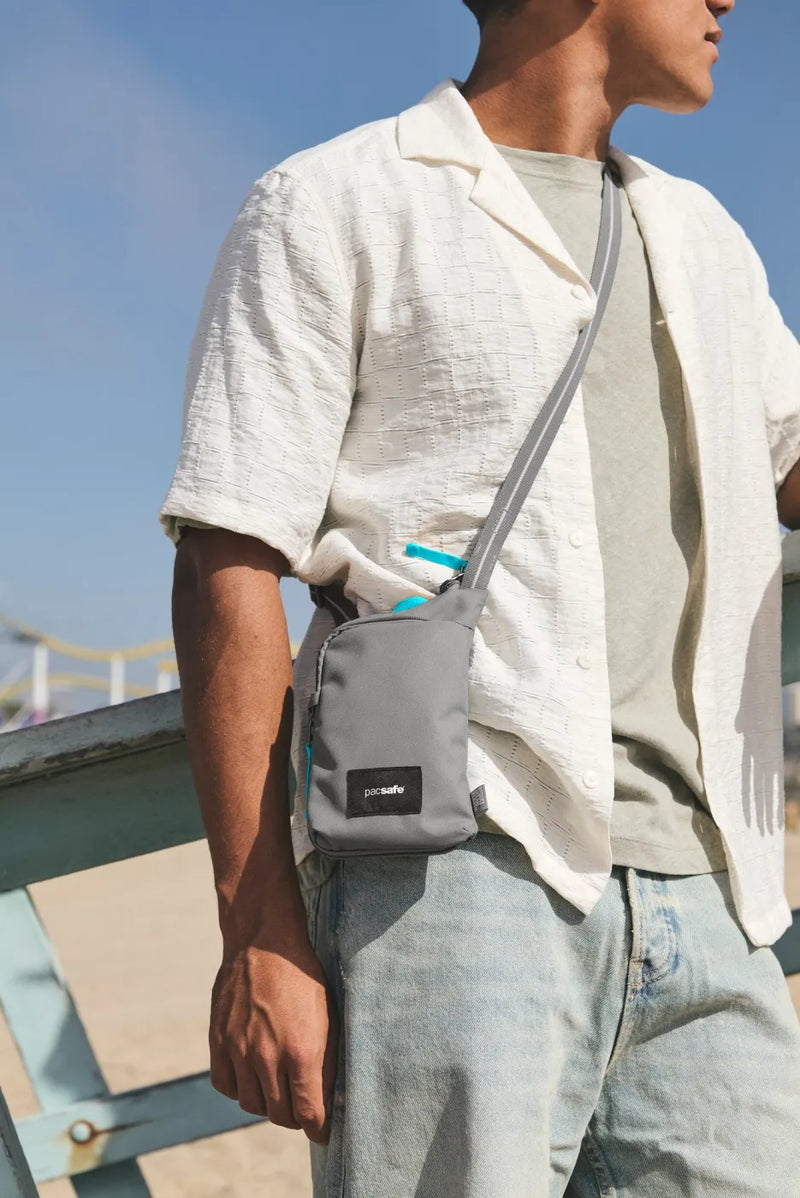 Pacsafe Go Anti-Theft Tech Crossbody Bag in Stone, worn crossbody at the hip by a man in a cream shirt at the beach