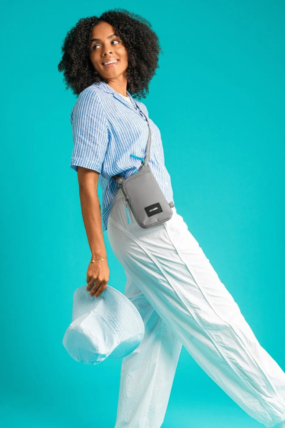 Pacsafe Go Anti-Theft Tech Crossbody Bag in Stone, worn crossbody by a woman in a striped shirt against a teal backdrop