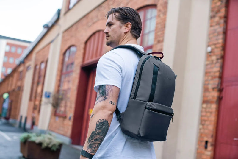 Pacsafe Metrosafe X Anti-Theft 20L 16" Laptop Backpack in Slate, worn on both shoulders by a man walking past a brick warehouse
