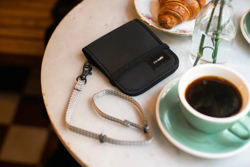 Pacsafe RFIDsafe V100 - Anti-theft RFID Blocking Bi-Fold Wallet in Black, on a cafe table beside a coffee and a croissant