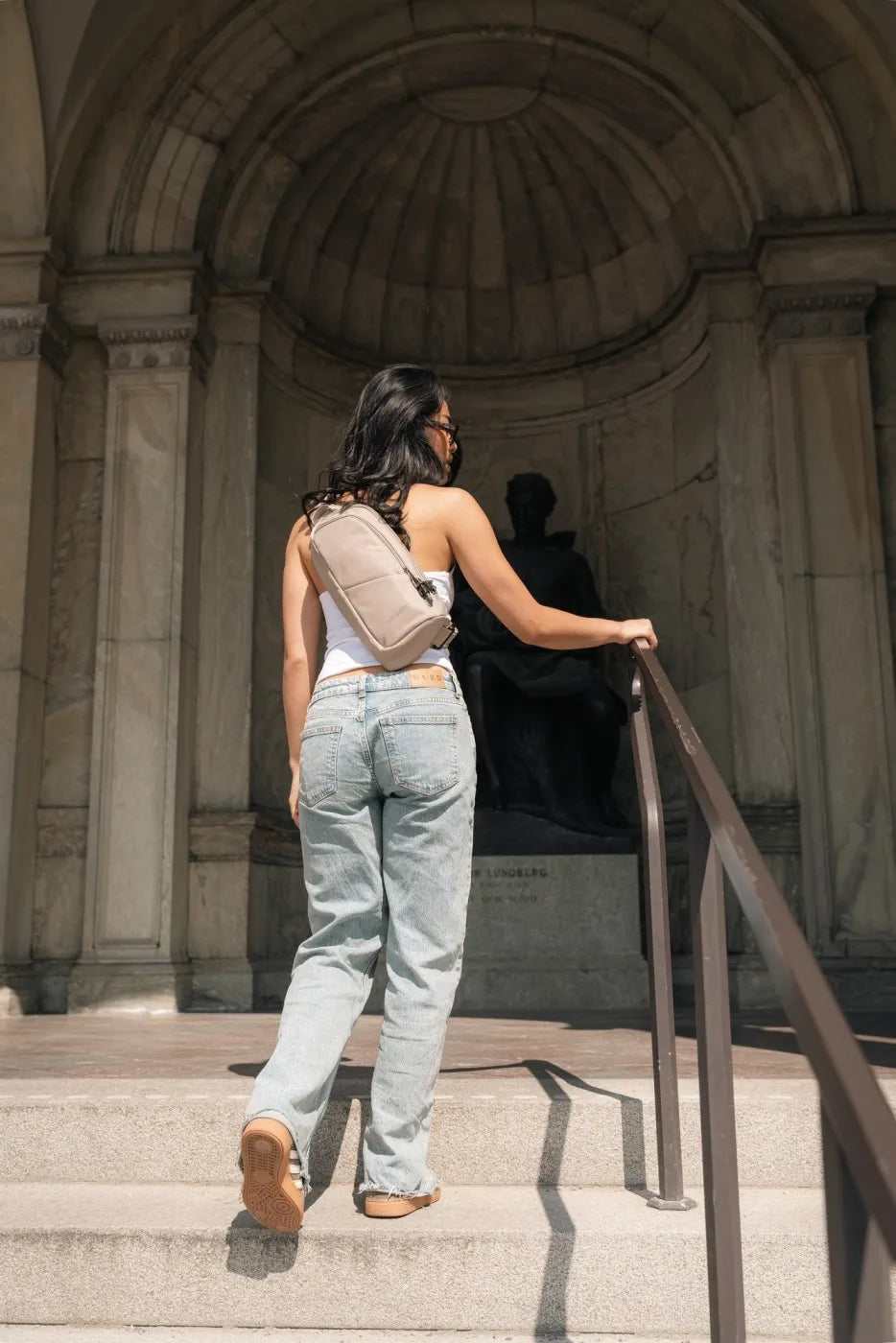 Pacsafe V Action Anti-Theft Sling Pack in Beige, worn crossbody at the back by a woman walking up steps to a monument