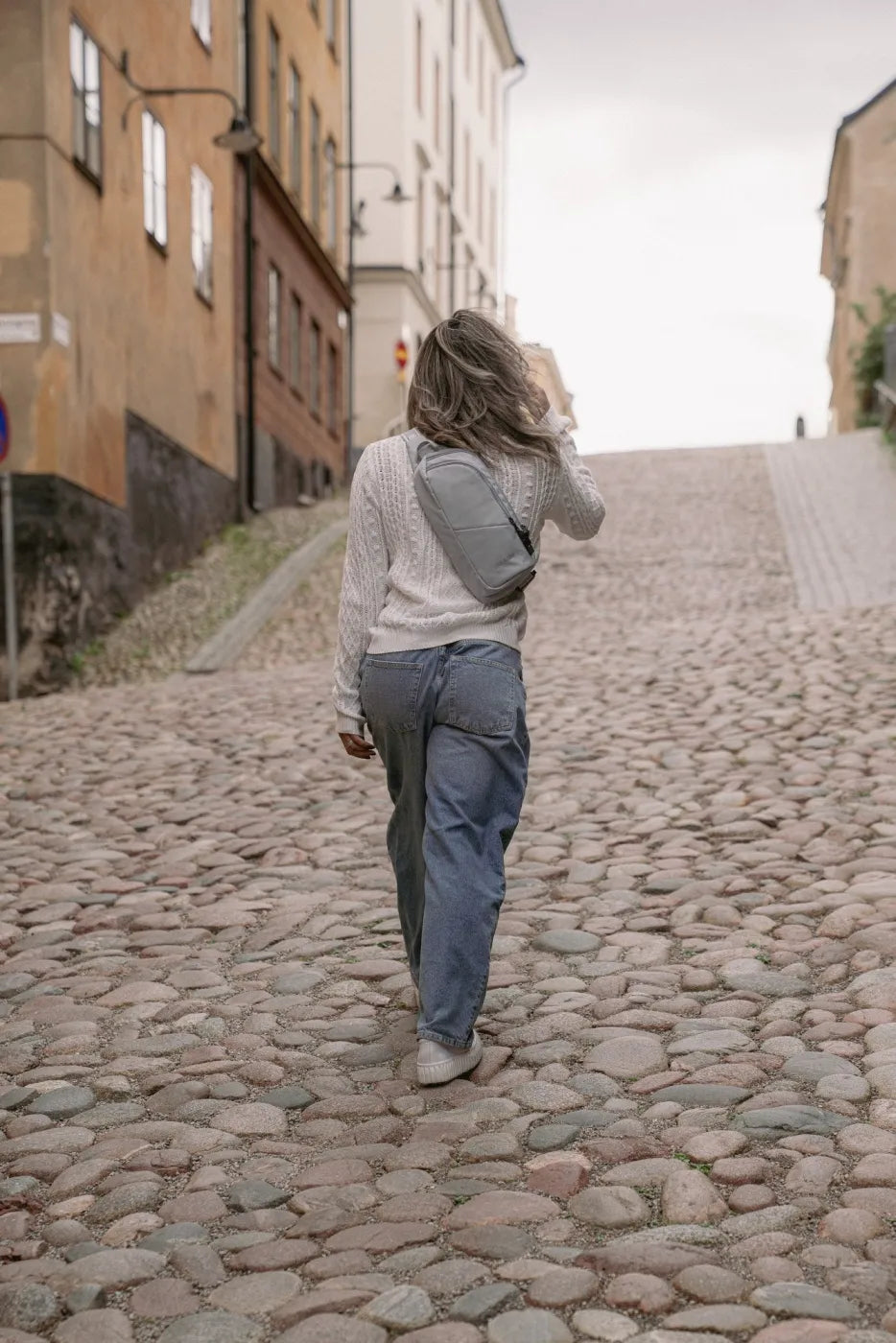Pacsafe V Action Anti-Theft Sling Pack in Digital Grey, worn crossbody by a woman walking up a cobbled hill