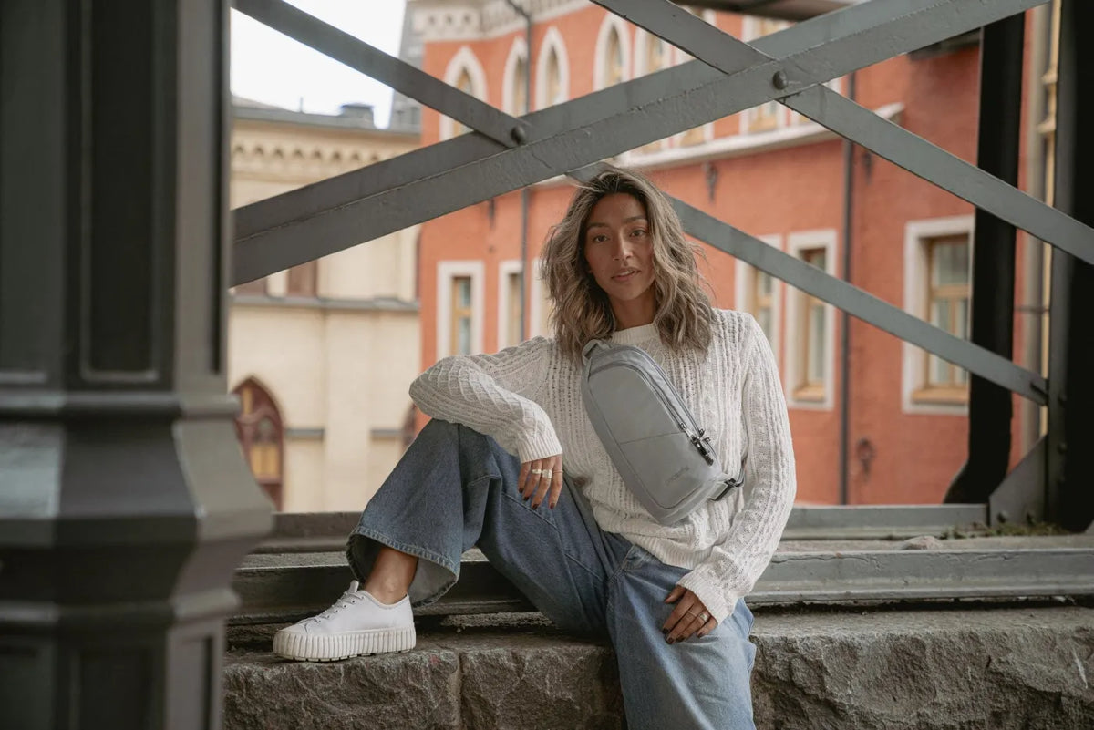Pacsafe V Action Anti-Theft Sling Pack in Digital Grey, worn crossbody by a woman seated on stone steps in front of a red building
