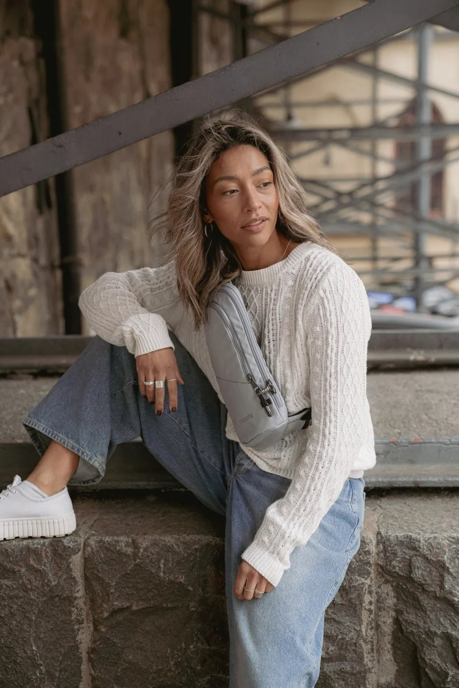 Pacsafe V Action Anti-Theft Sling Pack in Digital Grey, worn crossbody by a woman sitting on a stone ledge beneath a bridge