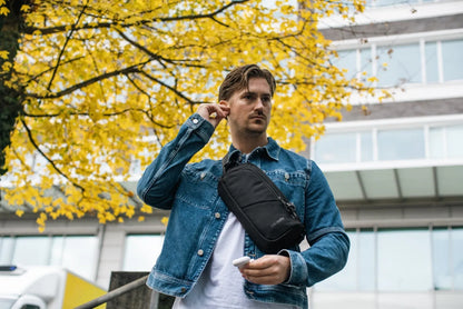 Pacsafe V Action Anti-Theft Sling Pack in Jet Black, worn crossbody by a man under an autumn tree, earbud in hand