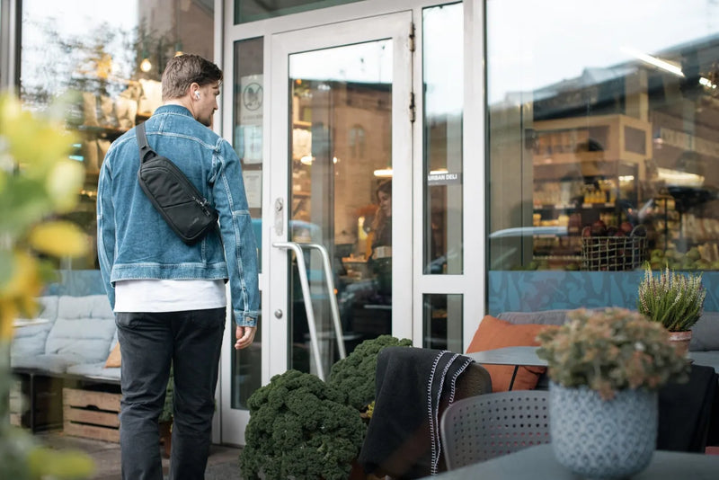 Pacsafe V Action Anti-Theft Sling Pack in Jet Black, worn crossbody at the back by a man entering a shop, seen from behind