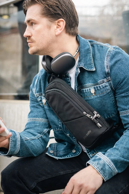 Pacsafe V Action Anti-Theft Sling Pack in Jet Black, worn crossbody by a seated man with headphones round his neck