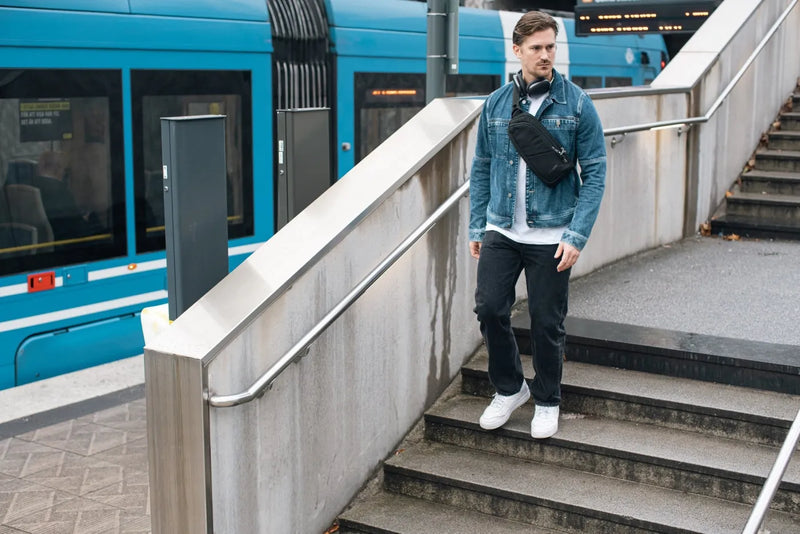 Pacsafe V Action Anti-Theft Sling Pack in Jet Black, worn crossbody by a man walking down station steps past a tram