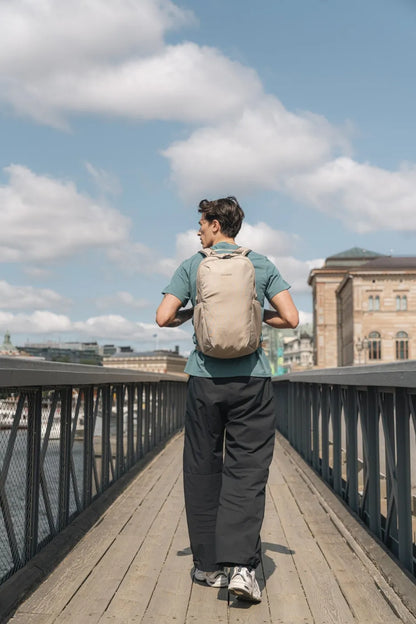 Pacsafe V Active 24L Anti-Theft 13" Laptop Backpack in Beige, worn on both shoulders by a man walking across a timber bridge