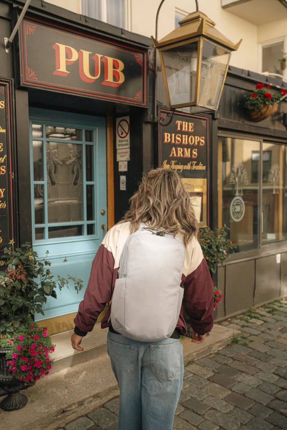 Pacsafe V Active 24L Anti-Theft 13" Laptop Backpack in Digital Grey, worn on both shoulders by a woman walking past a corner pub