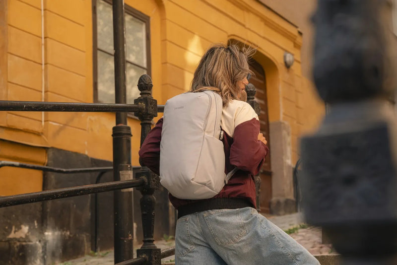 Pacsafe V Active 24L Anti-Theft 13" Laptop Backpack in Digital Grey, worn on both shoulders by a woman sitting on stone steps in an old town street