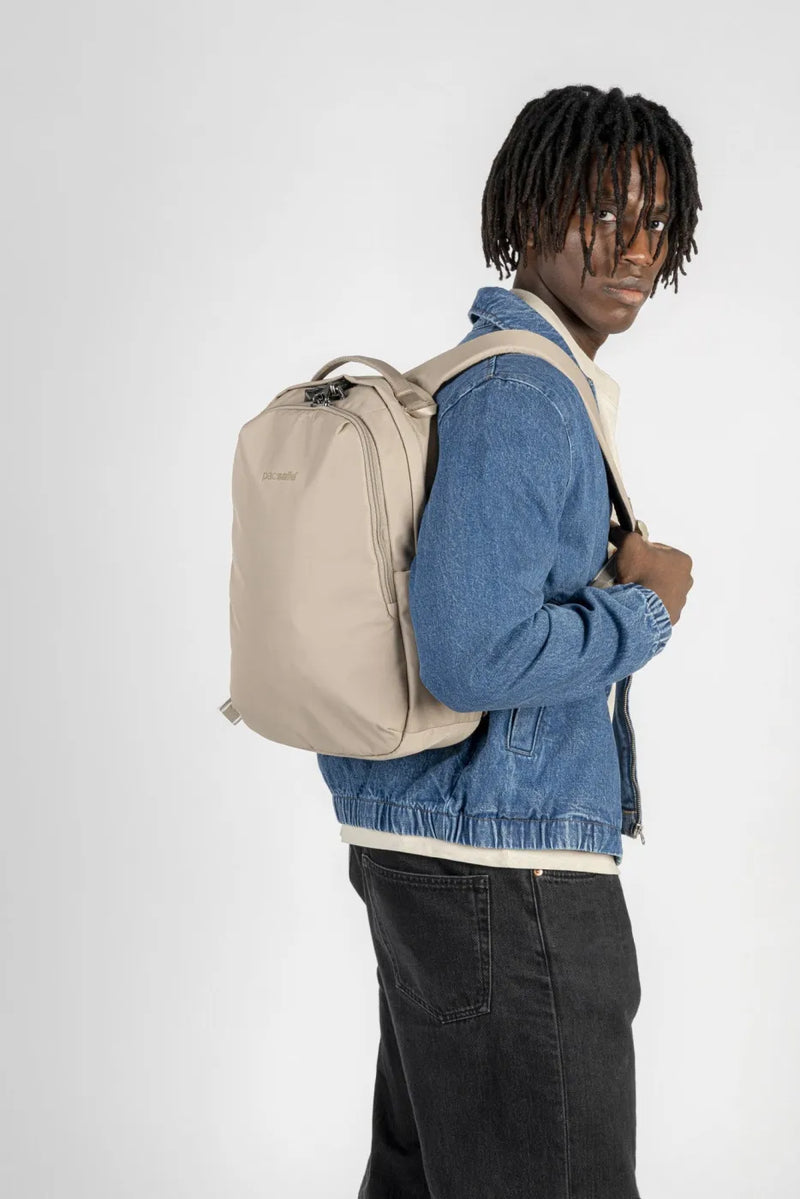 Pacsafe V All-Around 16L Anti-Theft 13" Laptop Backpack in Beige, worn on both shoulders by a man in a denim jacket, looking over his shoulder