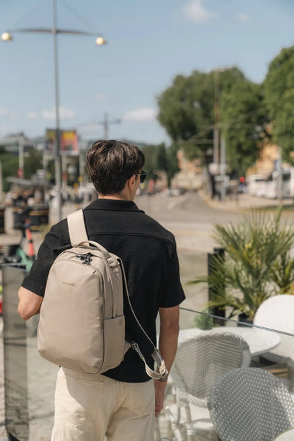 Pacsafe V All-Around 16L Anti-Theft 13" Laptop Backpack in Beige, worn on one shoulder by a man walking past a cafe terrace