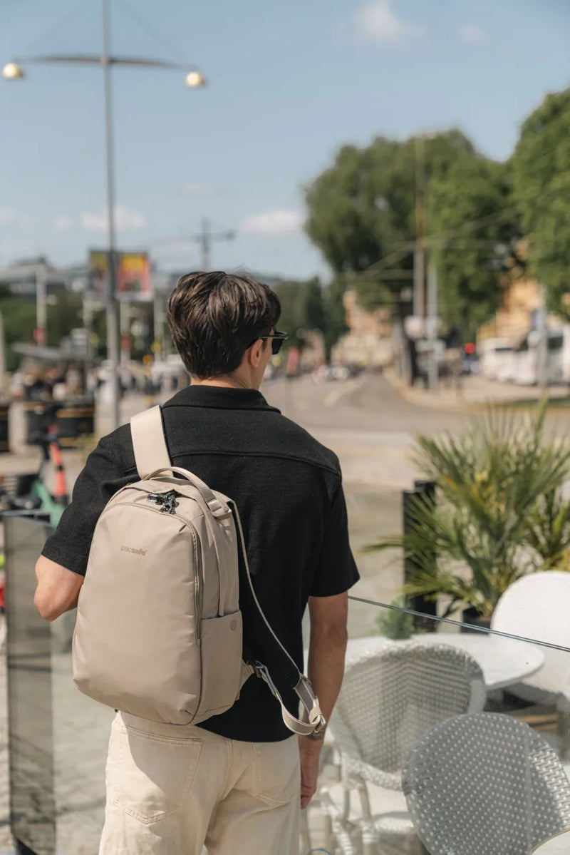 Pacsafe V All-Around 16L Anti-Theft 13" Laptop Backpack in Beige, worn on one shoulder by a man walking past a cafe terrace