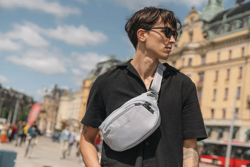 Pacsafe V Anti-Theft Hip Pack in Beige, worn crossbody at chest height by a man on a sunny city waterfront