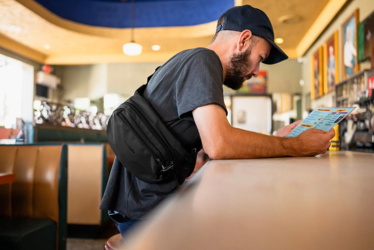 Pacsafe V Anti-Theft Hip Pack in Jet Black, worn crossbody by a man reading a menu at a diner counter