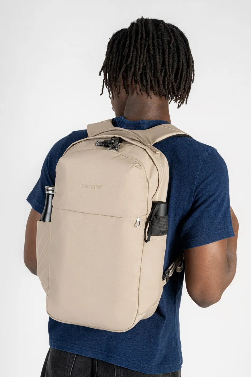 Pacsafe V City 20L Anti-Theft 16" Laptop Backpack in Beige, worn on both shoulders by a man in a navy tee, water bottle in the side pocket