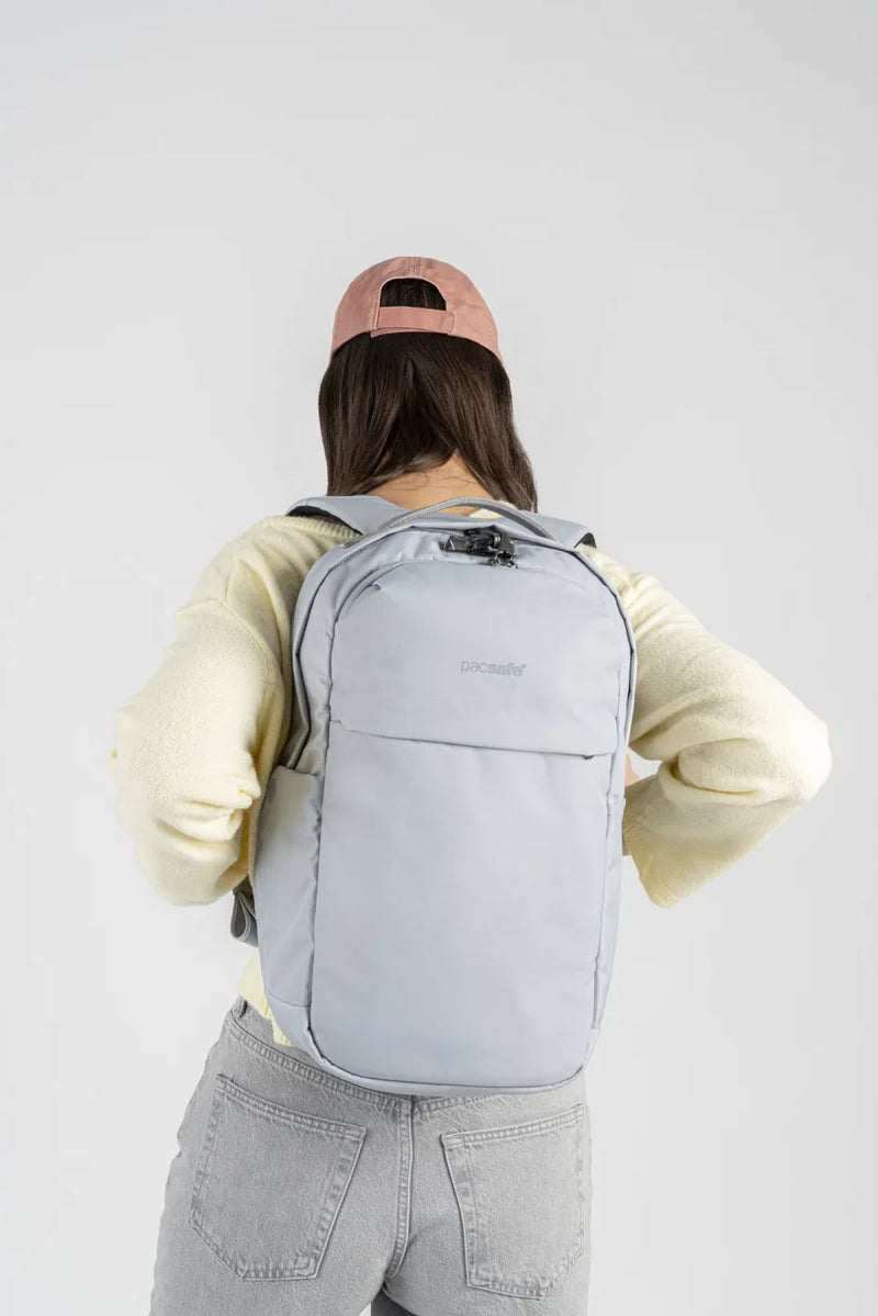 Pacsafe V City 20L Anti-Theft 16" Laptop Backpack in Digital Grey, worn on both shoulders by a woman in a pink cap, seen from behind