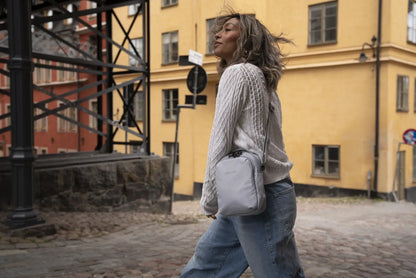 Pacsafe V Companion Anti-Theft Crossbody Bag in Digital Grey, worn crossbody by a woman walking a cobbled street past a yellow building