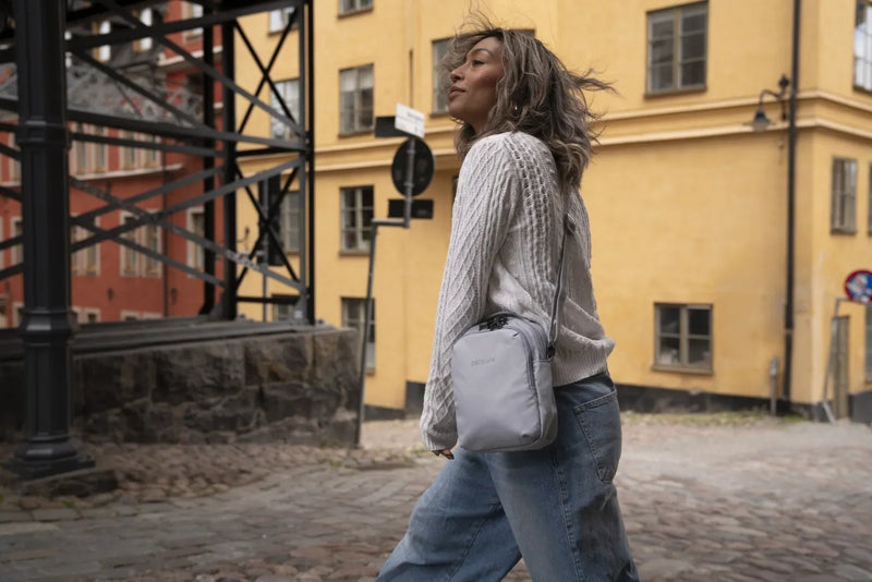 Pacsafe V Companion Anti-Theft Crossbody Bag in Digital Grey, worn crossbody by a woman walking a cobbled street past a yellow building