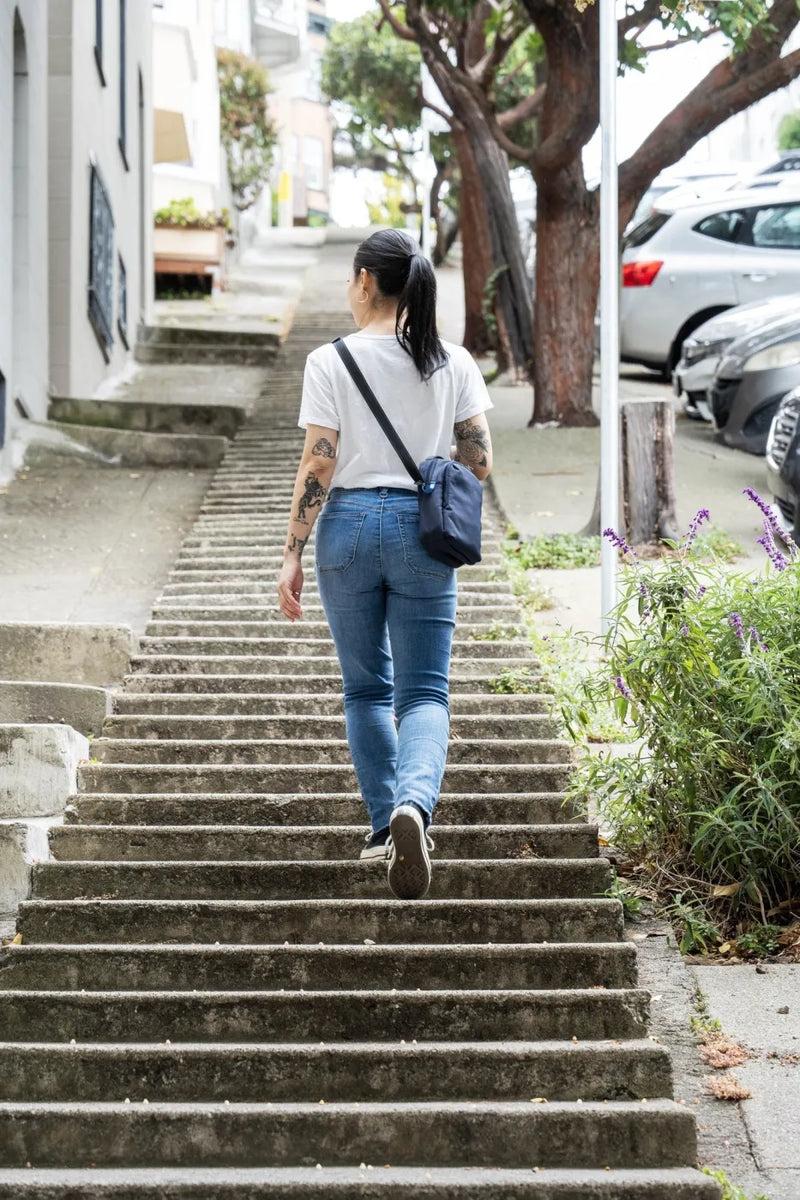Pacsafe V Companion Anti-Theft Crossbody Bag in Ocean, worn crossbody by a woman climbing a flight of city steps