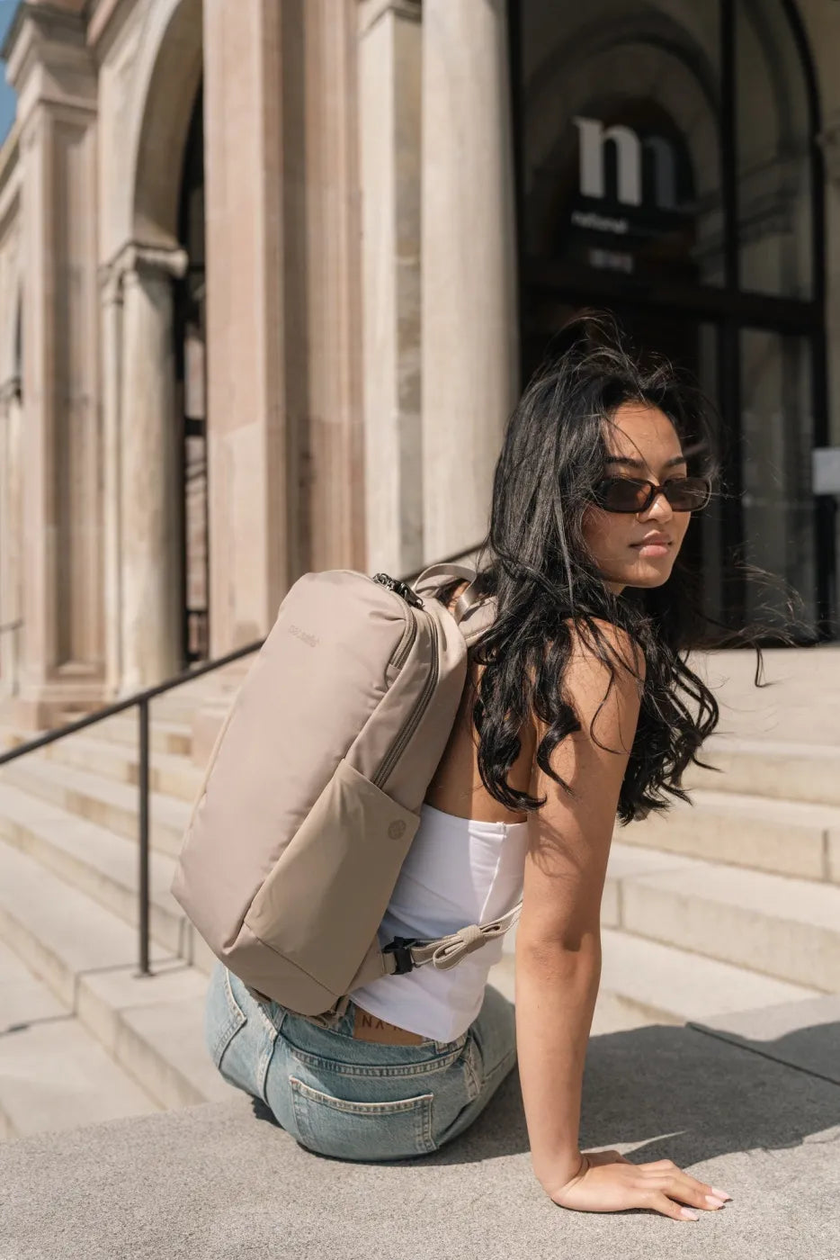 Pacsafe V Tech 12L Anti-Theft Backpack in Beige, worn on one shoulder by a woman crouching on stone steps outside a museum