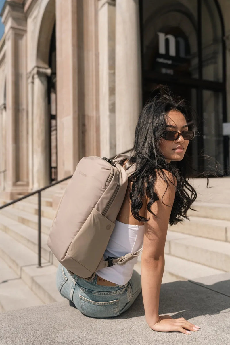 Pacsafe V Tech 12L Anti-Theft Backpack in Beige, worn on one shoulder by a woman crouching on stone steps outside a museum