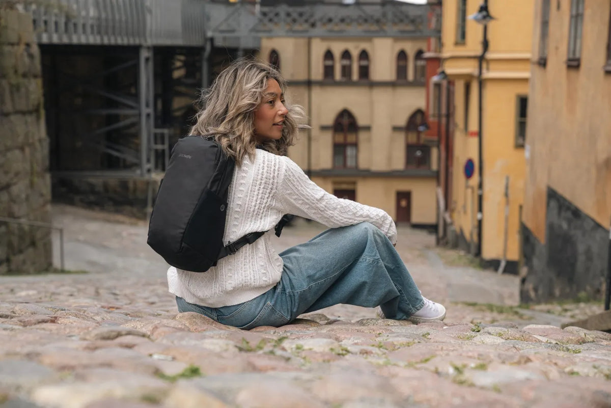 Pacsafe V Tech 12L Anti-Theft Backpack in Black, worn on both shoulders by a woman sitting on cobbles in an old town street