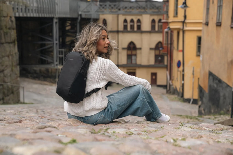 Pacsafe V Tech 12L Anti-Theft Backpack in Black, worn on both shoulders by a woman sitting on cobbles in an old town street