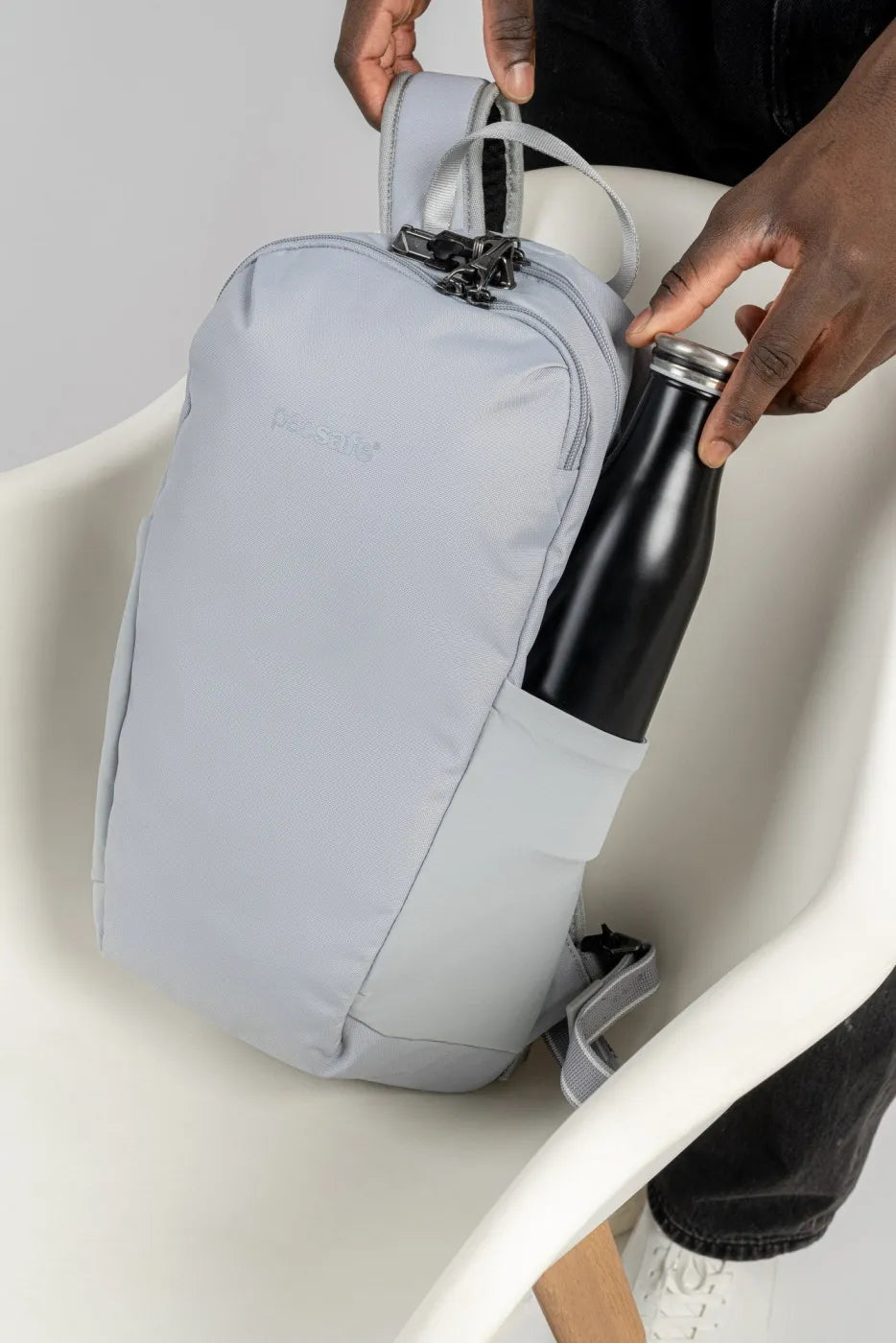 Pacsafe V Tech 12L Anti-Theft Backpack in Digital Grey, a water bottle being slid into the side pocket while the bag rests on a chair