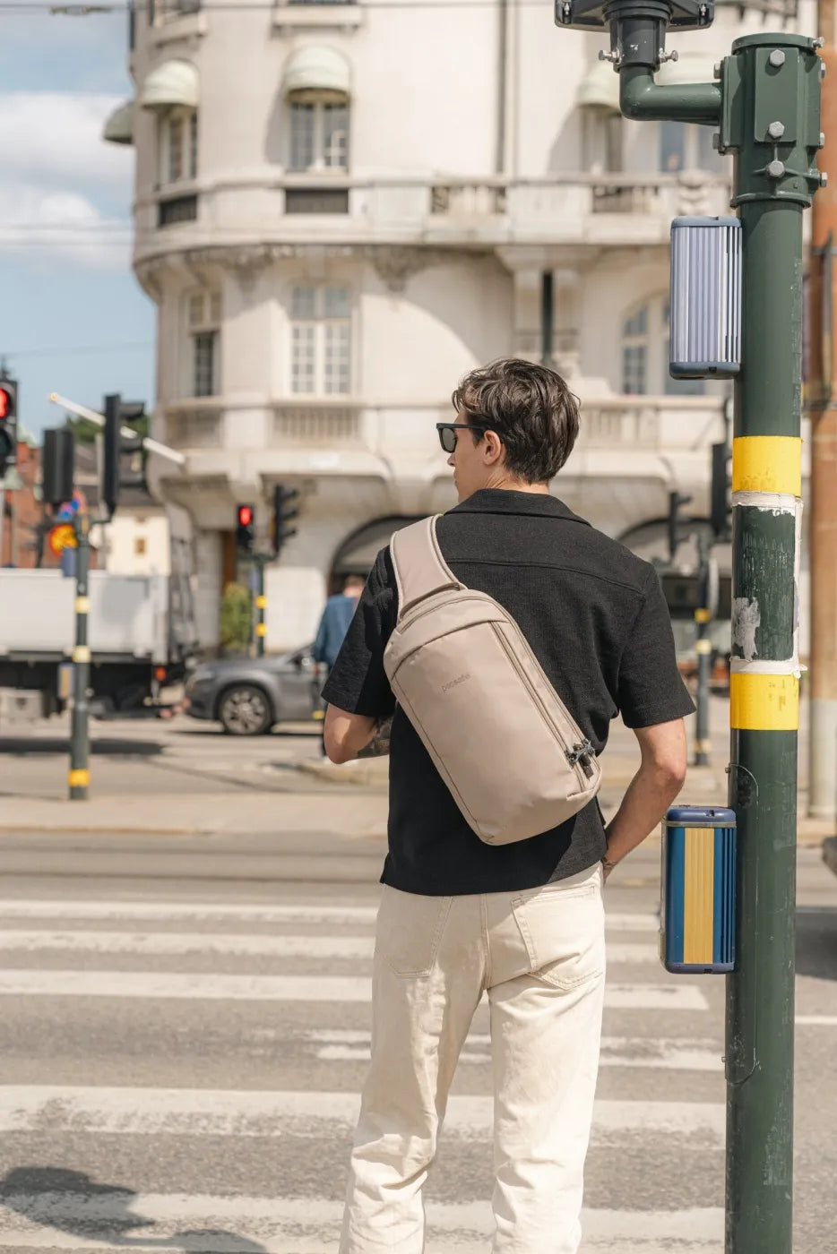 Pacsafe V Tech Anti-Theft 10L Sling Pack in Beige, worn on one shoulder by a man waiting at a pedestrian crossing