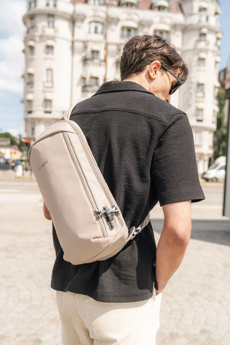 Pacsafe V Tech Anti-Theft 10L Sling Pack in Beige, worn across the back by a man in a dark polo shirt on a city street
