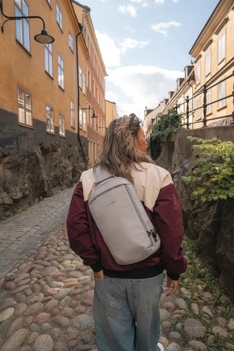 Pacsafe V Tech Anti-Theft 10L Sling Pack in Digital Grey, worn on one shoulder by a woman walking up a cobbled lane, seen from behind