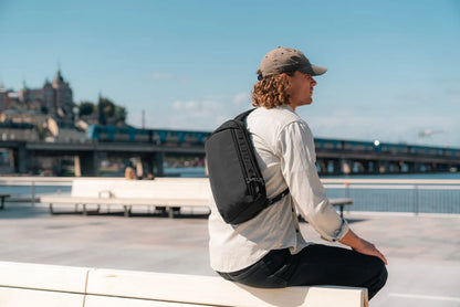 Pacsafe V Tech Anti-Theft 10L Sling Pack in Jet Black, worn across the chest by a man sitting on a quayside wall