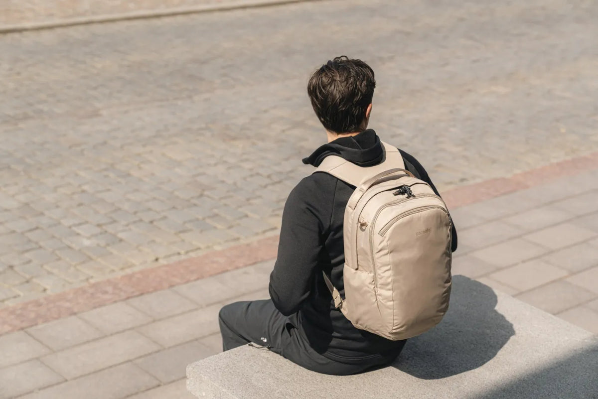 Pacsafe V Tour 26L Anti-Theft 16" Laptop Backpack in Beige, worn on both shoulders by a man sitting on a low wall in a city square