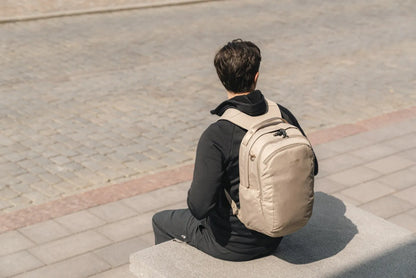 Pacsafe V Tour 26L Anti-Theft 16" Laptop Backpack in Beige, worn on both shoulders by a man sitting on a low wall in a city square