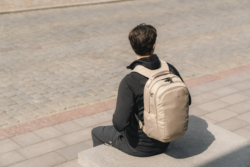 Pacsafe V Tour 26L Anti-Theft 16" Laptop Backpack in Beige, worn on both shoulders by a man sitting on a low wall in a city square