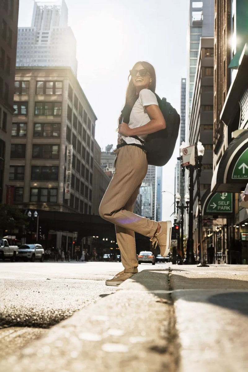 Pacsafe V Tour 26L Anti-Theft 16" Laptop Backpack in Ocean, worn on one shoulder by a woman jumping on a downtown pavement