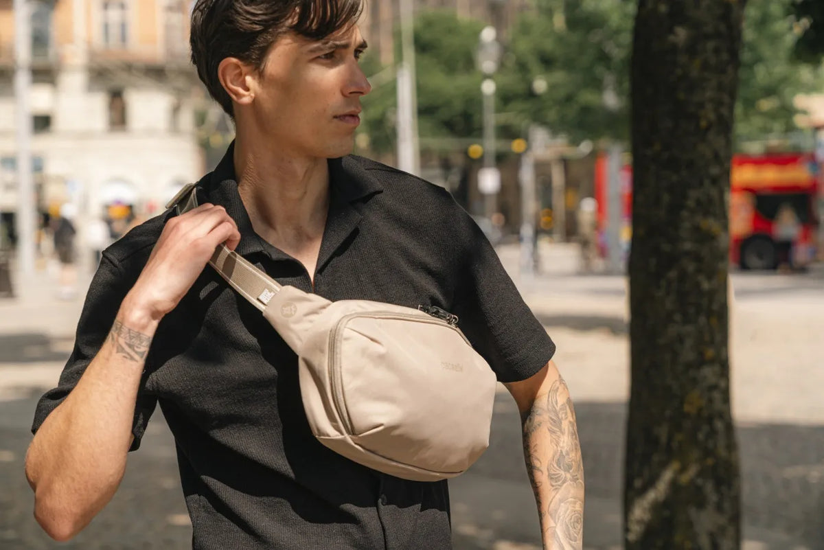 Pacsafe V Urban Anti-Theft Sling Pack in Beige, worn across the chest by a man in a black polo, strap in hand