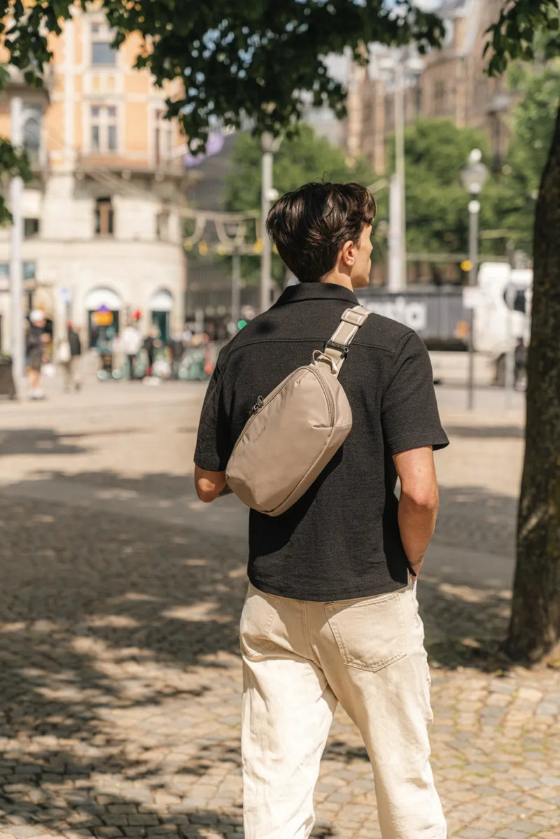 Pacsafe V Urban Anti-Theft Sling Pack in Beige, worn across the back by a man walking through a city square