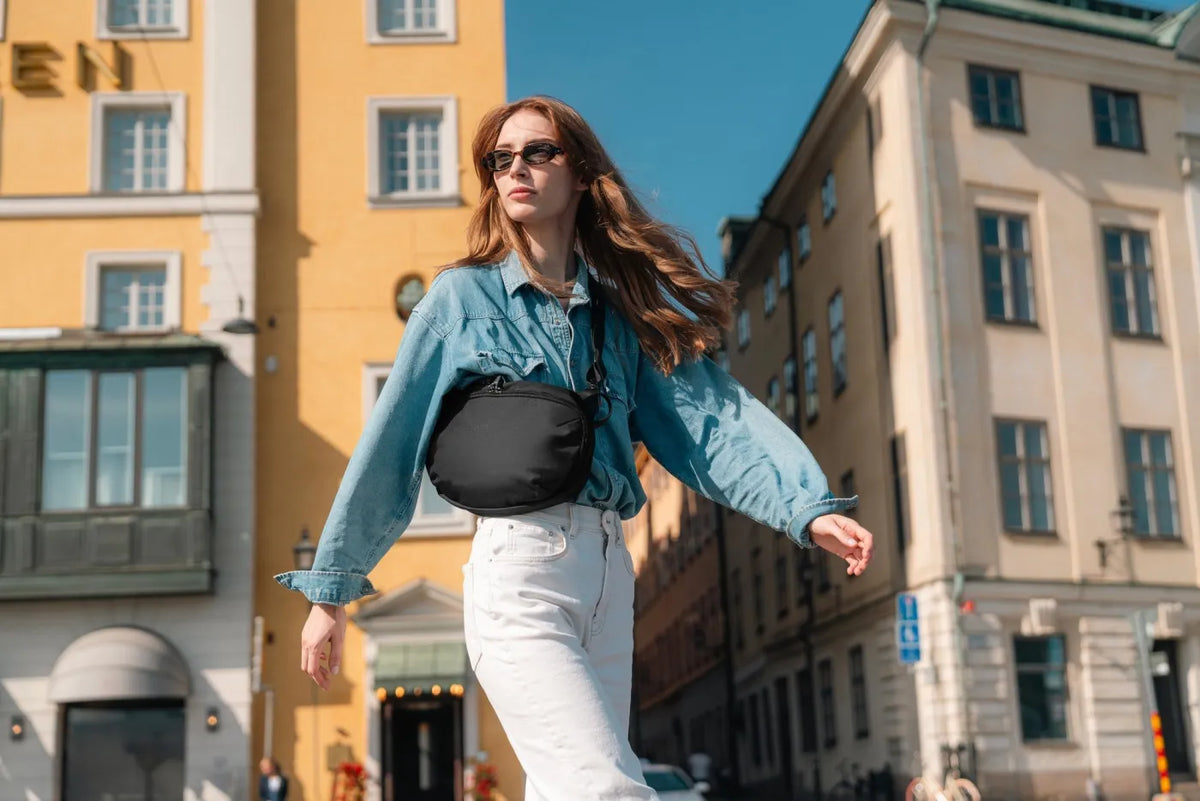 Pacsafe V Urban Anti-Theft Sling Pack in Black, worn at the waist by a woman walking a sunlit old town street