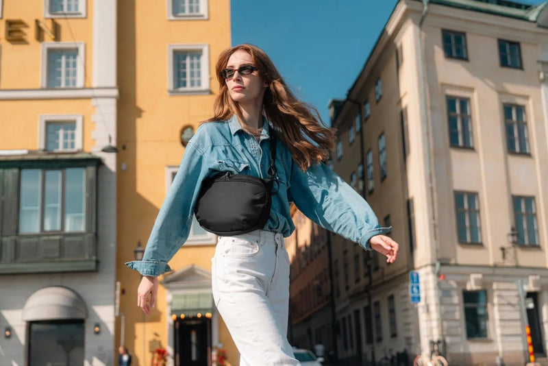 Pacsafe V Urban Anti-Theft Sling Pack in Black, worn at the waist by a woman walking a sunlit old town street