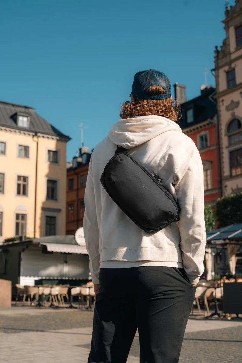Pacsafe V Urban Anti-Theft Sling Pack in Black, worn across the back over a cream hoodie, seen from behind