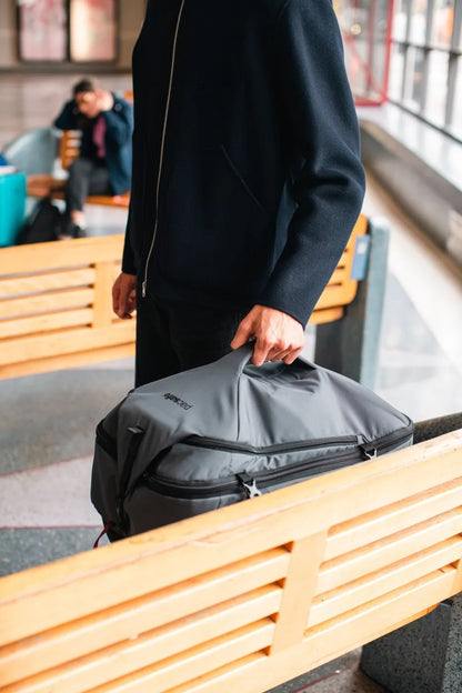 Pacsafe Venturesafe EXP45 Anti-Theft 45L Carry-on Travel Pack in Slate, carried by its top handle past a beachfront bench