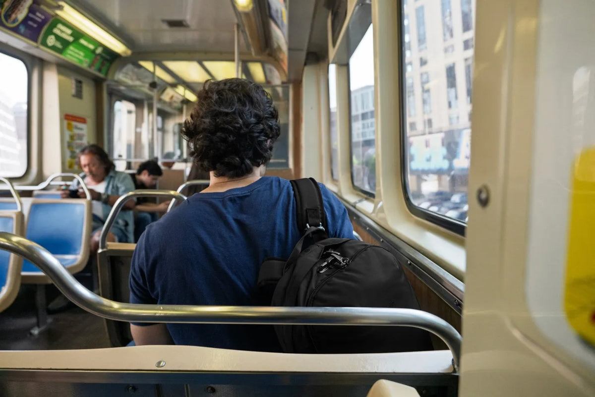 Pacsafe Vibe 25 - Anti-Theft 25L Laptop Daypack in Jet Black, resting on the seat beside a man riding a tram