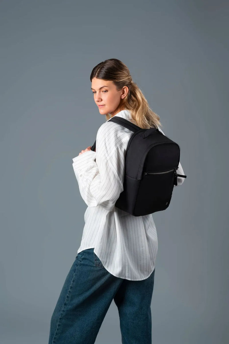 Pacsafe W 10L Anti-theft 13" Laptop Backpack in Black, worn on one shoulder by a woman looking back over her shoulder