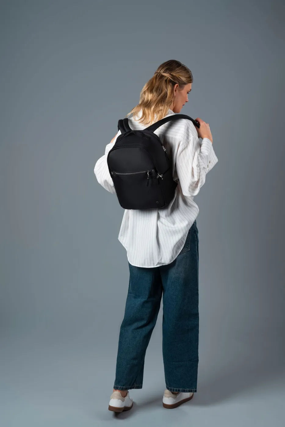 Pacsafe W 10L Anti-theft 13" Laptop Backpack in Black, swung onto the shoulders by a woman in a white shirt, seen from behind