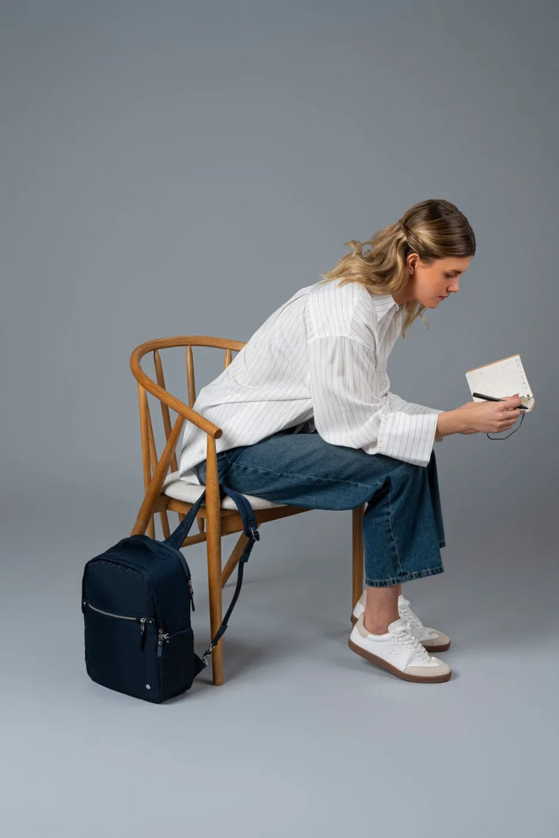 Pacsafe W 10L Anti-theft 13" Laptop Backpack in Navy, locked to the chair beside a woman writing in a notebook